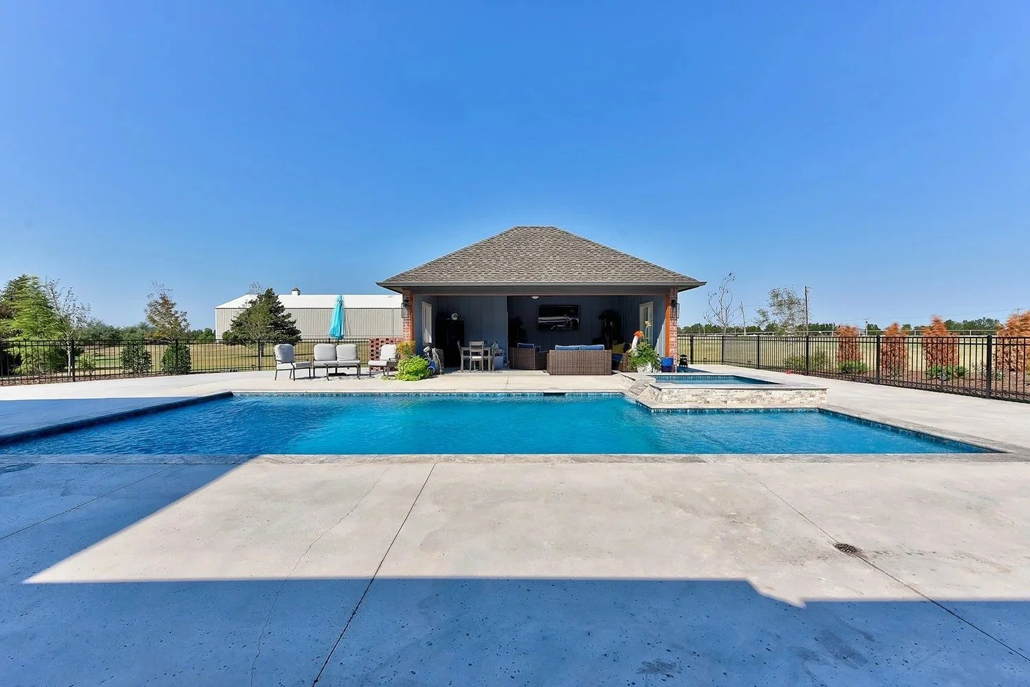 A backyard with a swimming pool, a pool house with outdoor furniture, lounge chairs, and a clear blue sky.