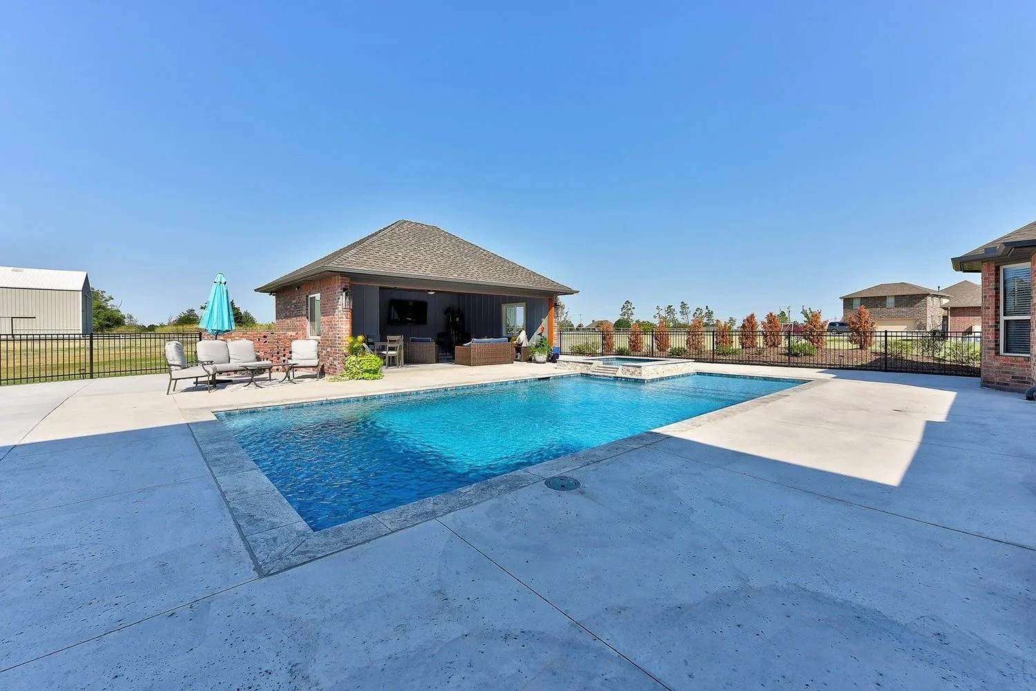 Backyard with in-ground swimming pool, outdoor seating area with chairs, table, and umbrella, adjacent to a covered patio with outdoor furniture, surrounded by a black metal fence.