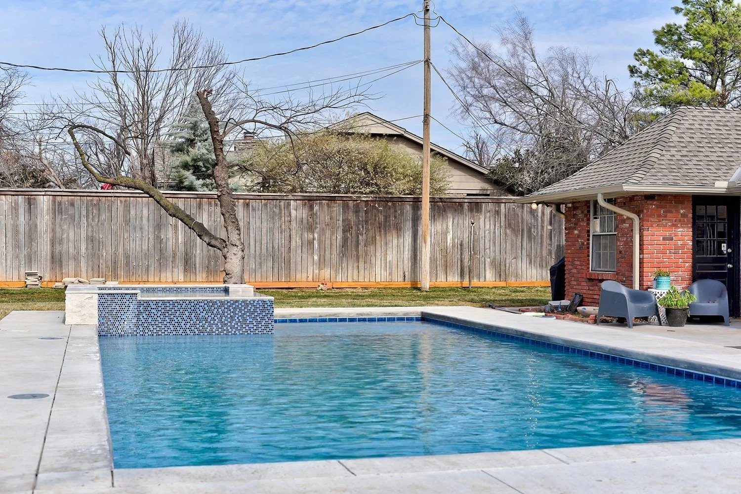 A backyard with a swimming pool, a leafless tree in a planter, a wooden fence, an electric pole, and a brick house with outdoor seating and potted plants.