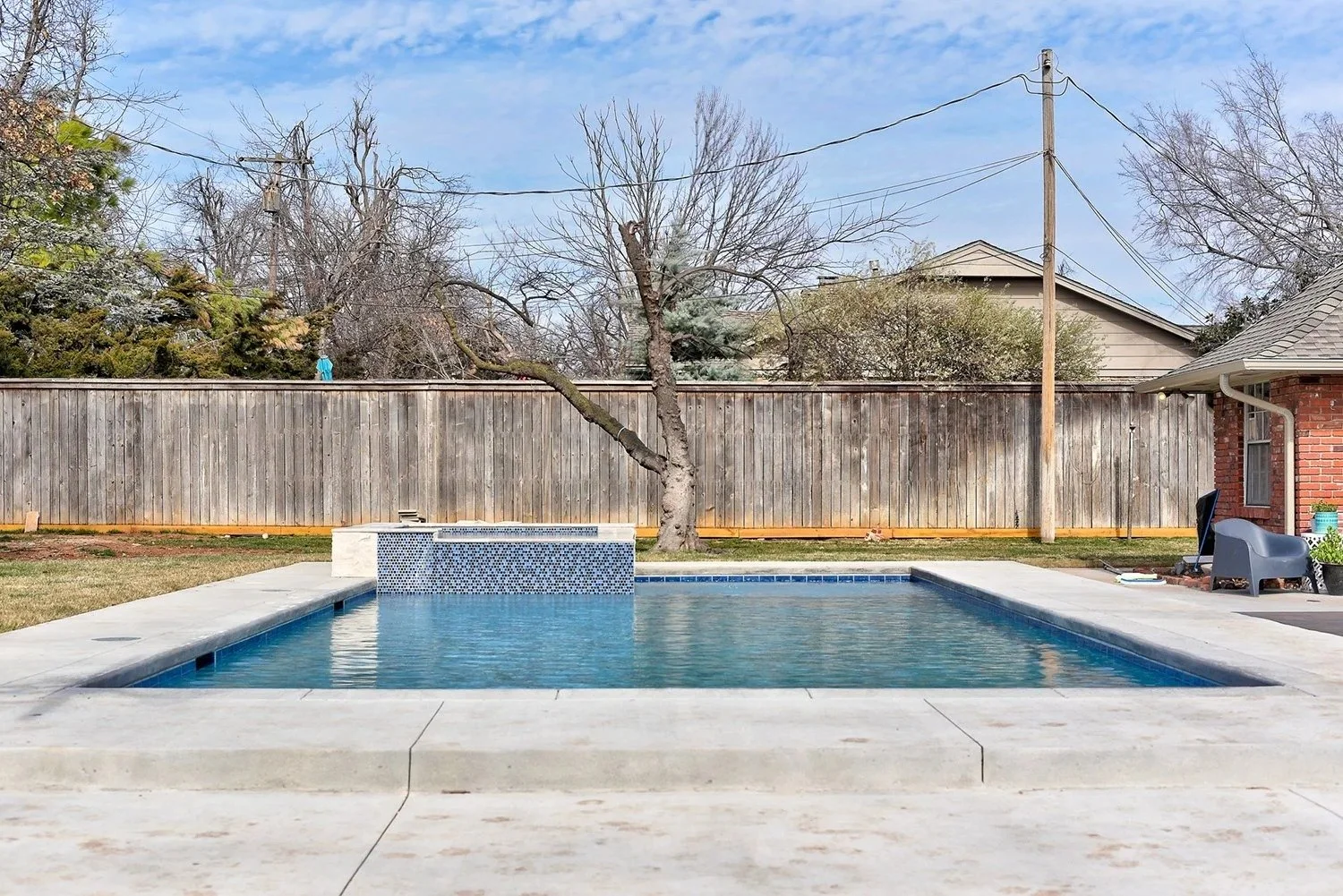 A backyard with a swimming pool, a wooden fence, a leafless tree, and a house with a brick wall and an outdoor chair.