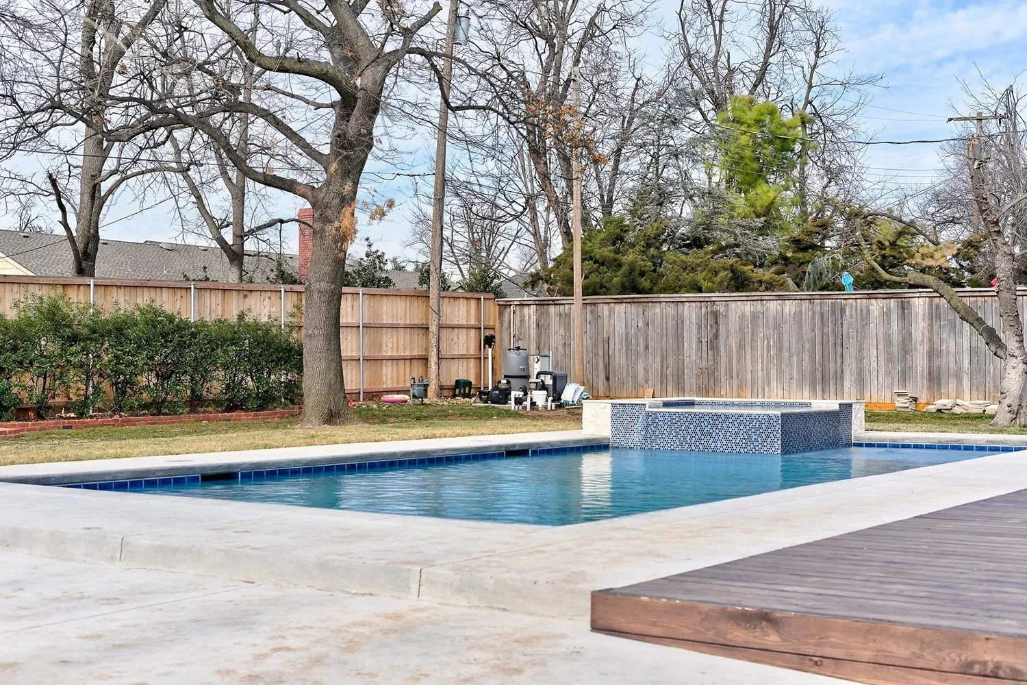A backyard with a swimming pool, surrounded by a concrete patio and a wooden deck. There are trees and a wooden fence in the background, along with some pool equipment.
