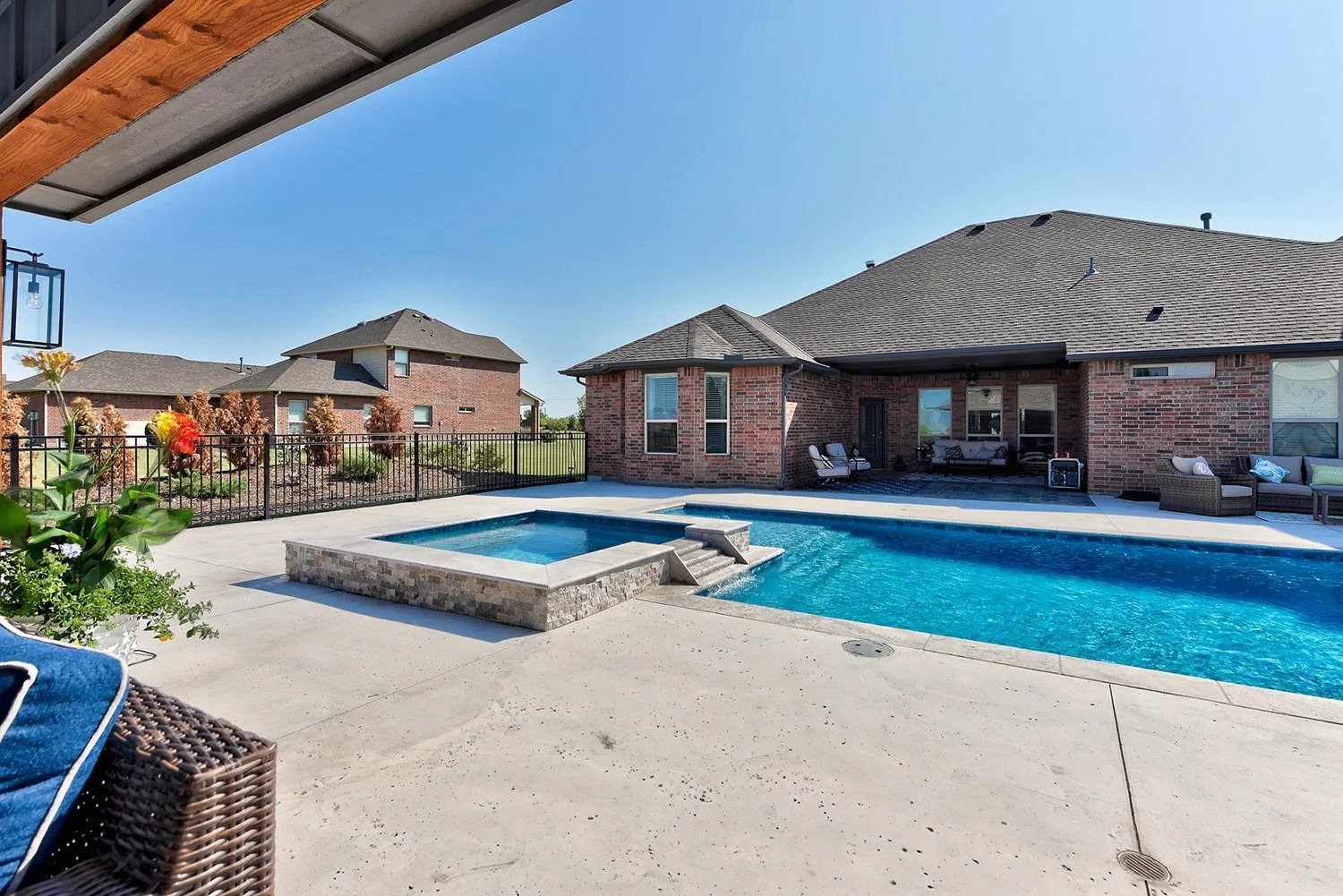Backyard with a swimming pool and hot tub, outdoor seating, brick house, fenced yard, and clear blue sky.