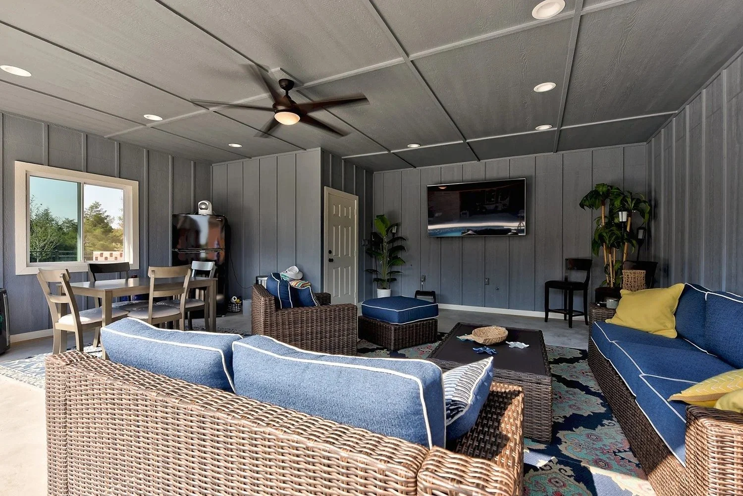 Living room with wicker furniture with blue cushions, a dark coffee table, a flat-screen TV on a wooden-paneled wall, potted plants, a ceiling fan, and a window showing greenery outside.