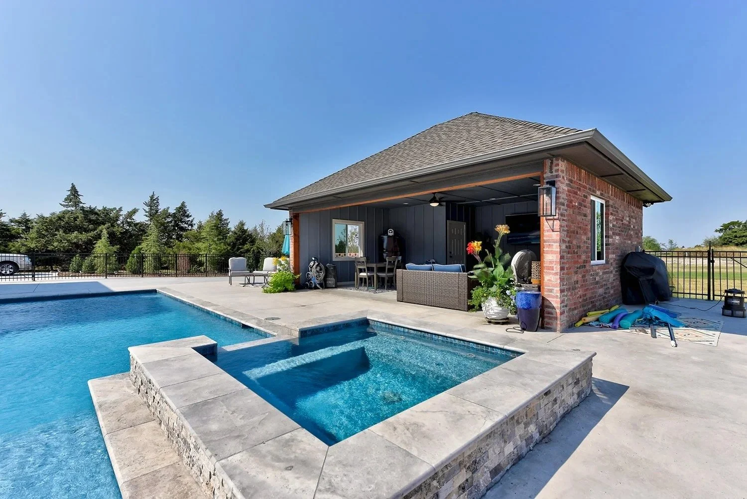 Backyard with a swimming pool and a pool house featuring outdoor furniture, potted plants, and pool toys on a sunny day.