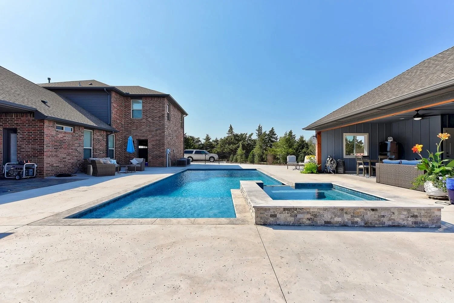 A backyard with a rectangular swimming pool and a hot tub, surrounded by patio furniture, including chairs, sofas, and umbrellas. There are two houses with brick and siding exteriors, trees in the background, and a clear blue sky.