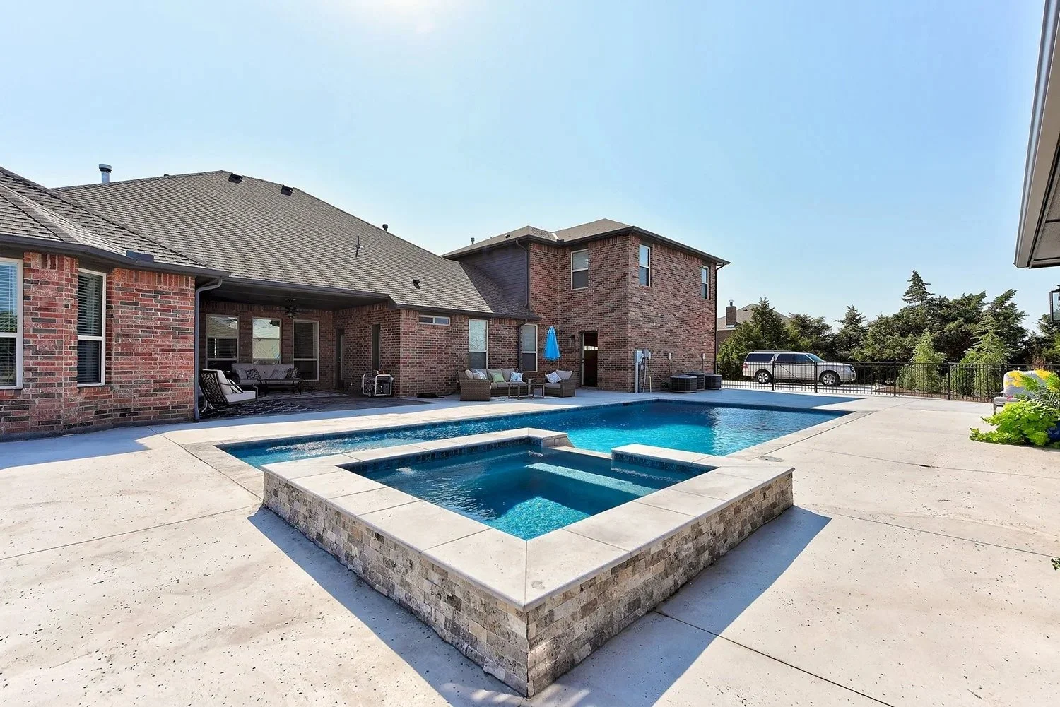 A backyard with a swimming pool and hot tub, surrounded by a concrete deck, with a brick house in the background, a parked car, and trees in the distance.