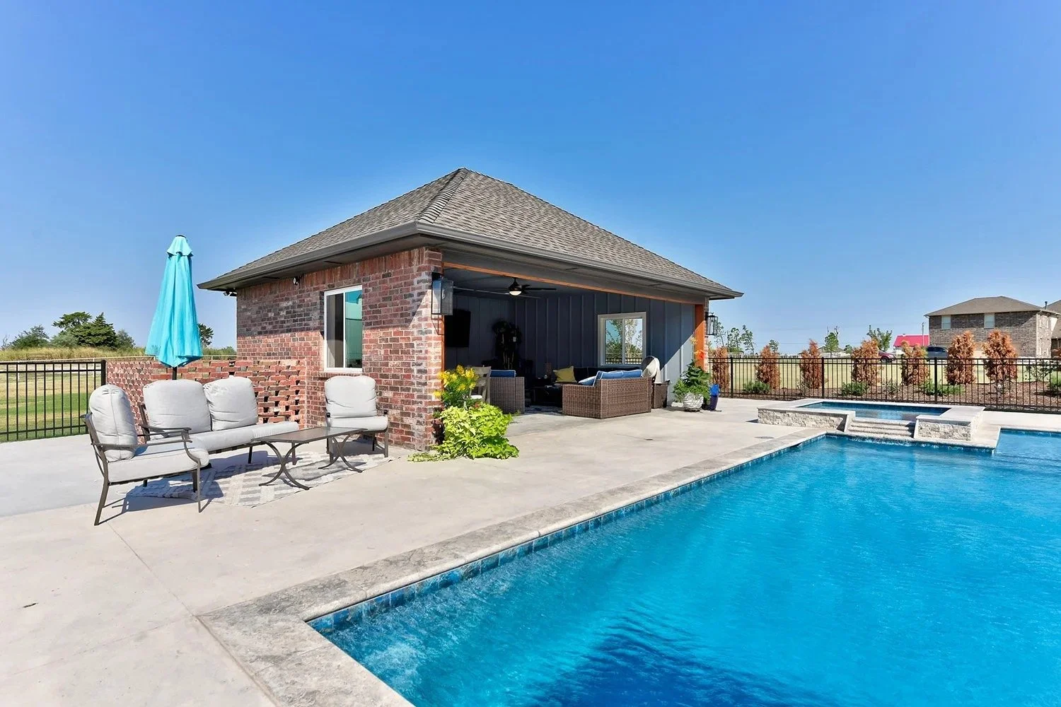 A backyard with a swimming pool, outdoor seating, and a brick pool house under a clear blue sky.
