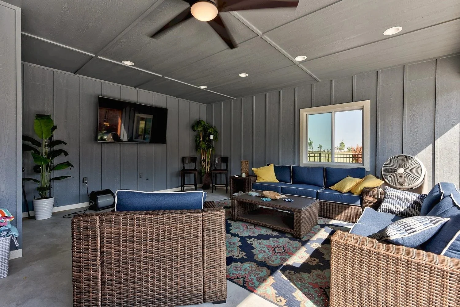 Living room with blue couch, wicker chairs, area rug, window, plants, TV, and ceiling fan.