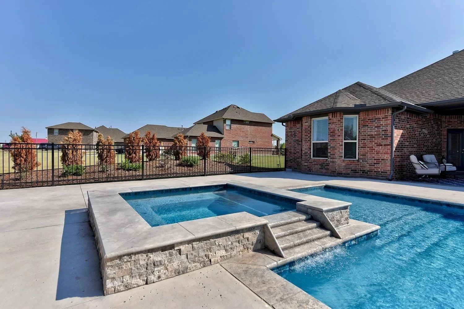 Residential backyard with a swimming pool and hot tub, brick house with patio chairs, fenced yard, neighboring houses, green lawn, and a clear blue sky.
