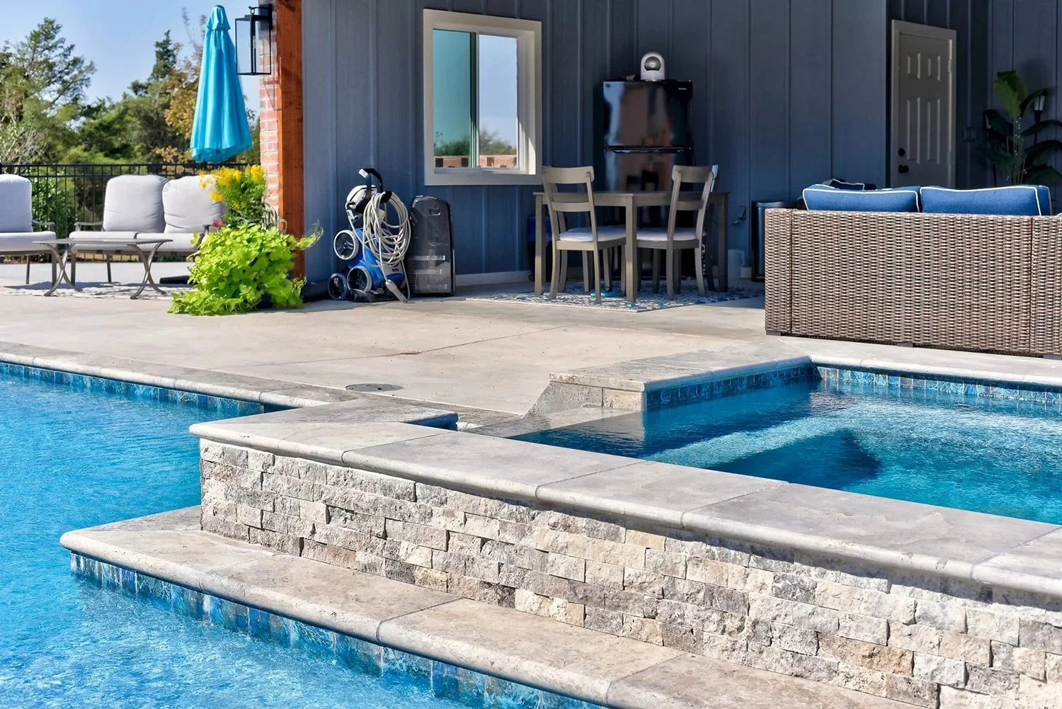 Backyard patio area with swimming pool, outdoor seating, patio furniture, green plants, and pool equipment against a blue house wall.