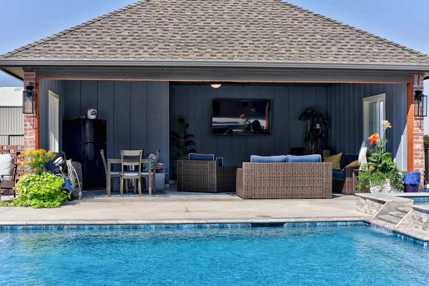 Backyard patio with outdoor seating, a pool, and a TV mounted on the wall of a house with gray siding and a shingled roof.