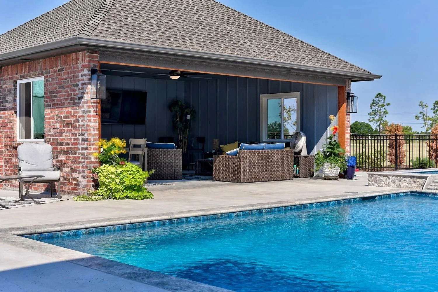 A backyard with a swimming pool and a covered patio. The patio has outdoor furniture, potted plants, and a television mounted on the wall. The area is surrounded by a black metal fence and trees in the background.