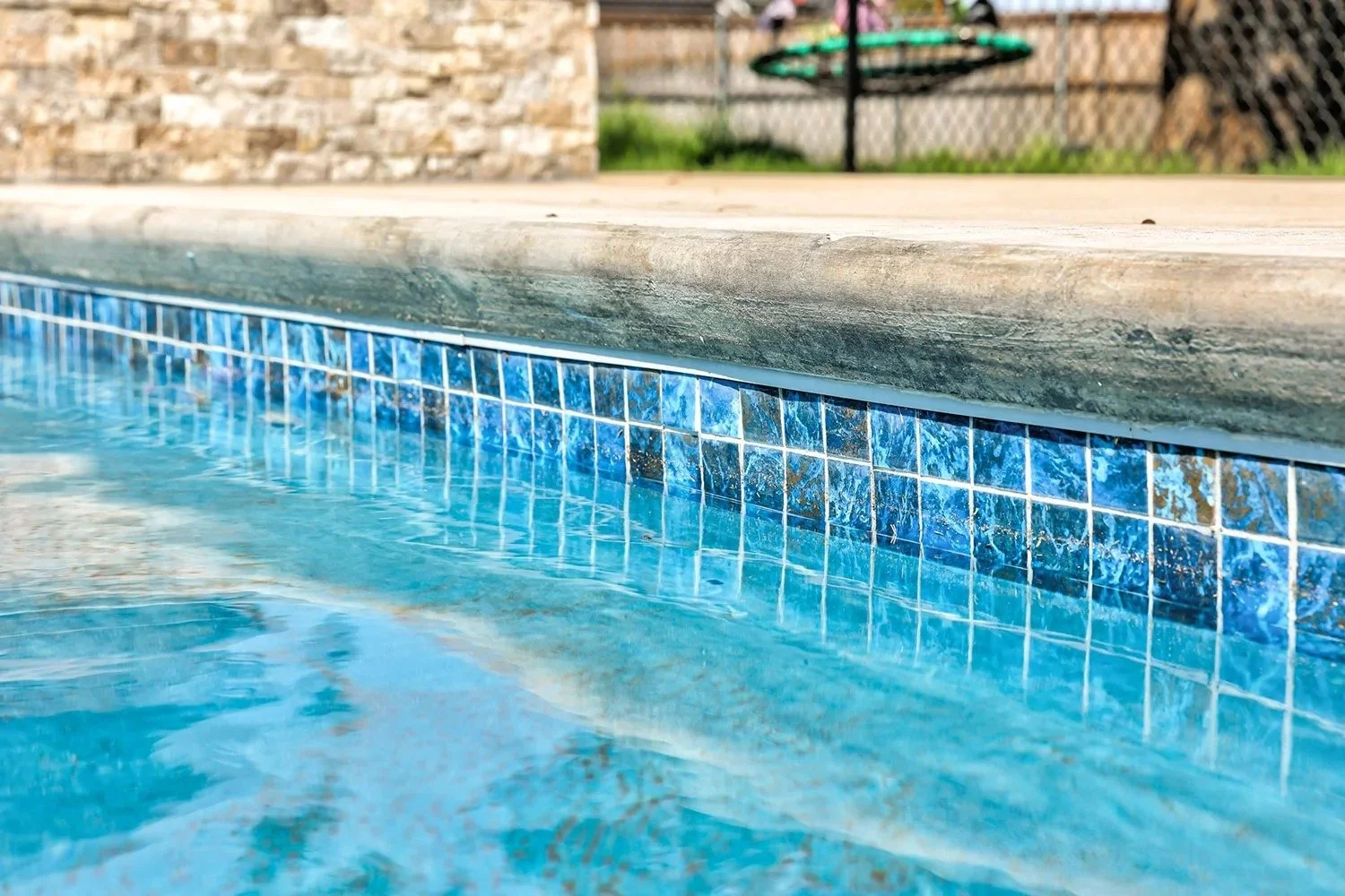 Close-up of a swimming pool's edge showing blue tiled border and water, with a concrete deck and a brick wall in the background.