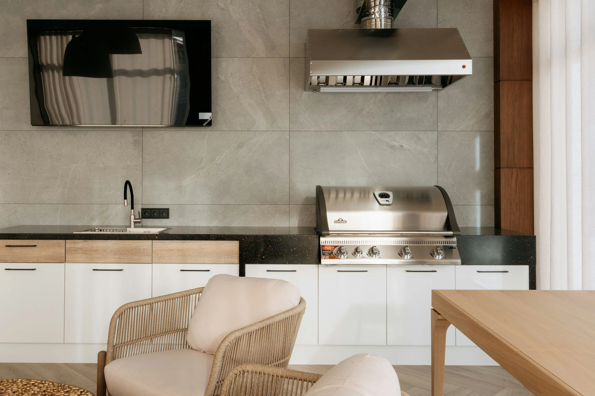 Modern kitchen with gray tiled wall, stainless steel oven, wooden cabinets, and a dining chair with a cushioned seat.