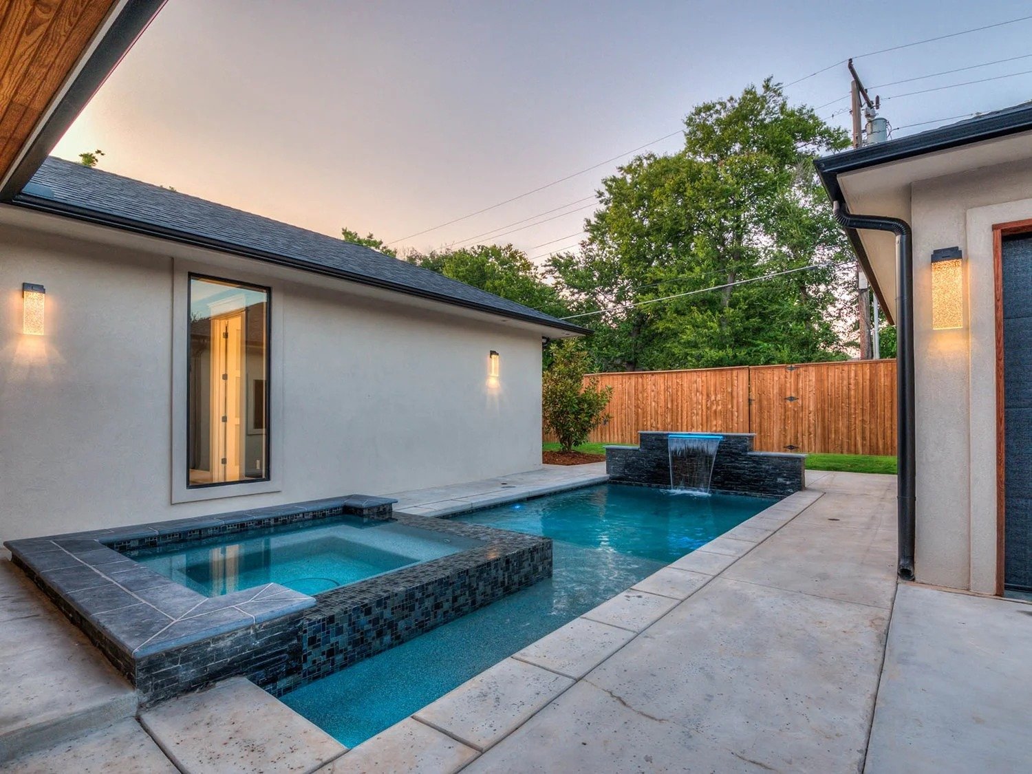 Backyard featuring a swimming pool with a waterfall, a hot tub, a wooden privacy fence, and exterior walls with lights at sunset.