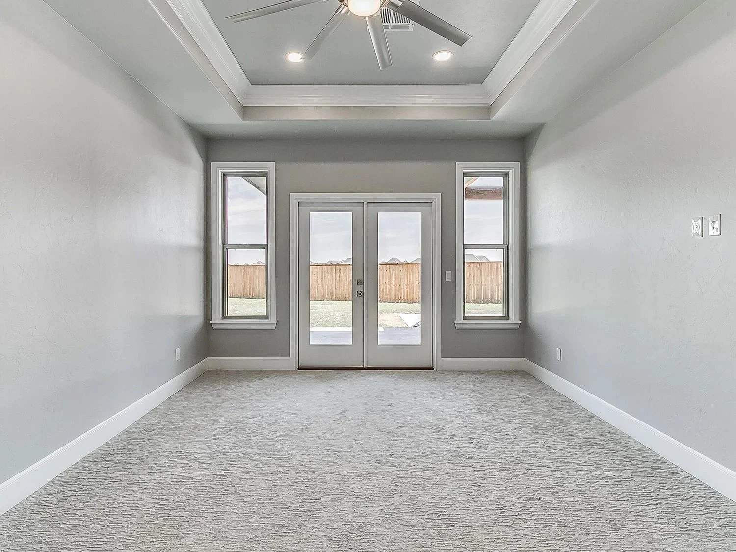 Empty room with light gray walls, white baseboards, a beige carpet, and a ceiling fan with lights. Double glass doors lead to an outdoor area with a wooden fence, and two narrow windows on either side of the door let in natural light.