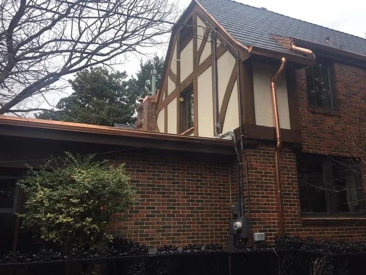 A brick house with a small addition featuring a half-timbered design on the upper part of the wall, copper gutters, and tired trees in the background.