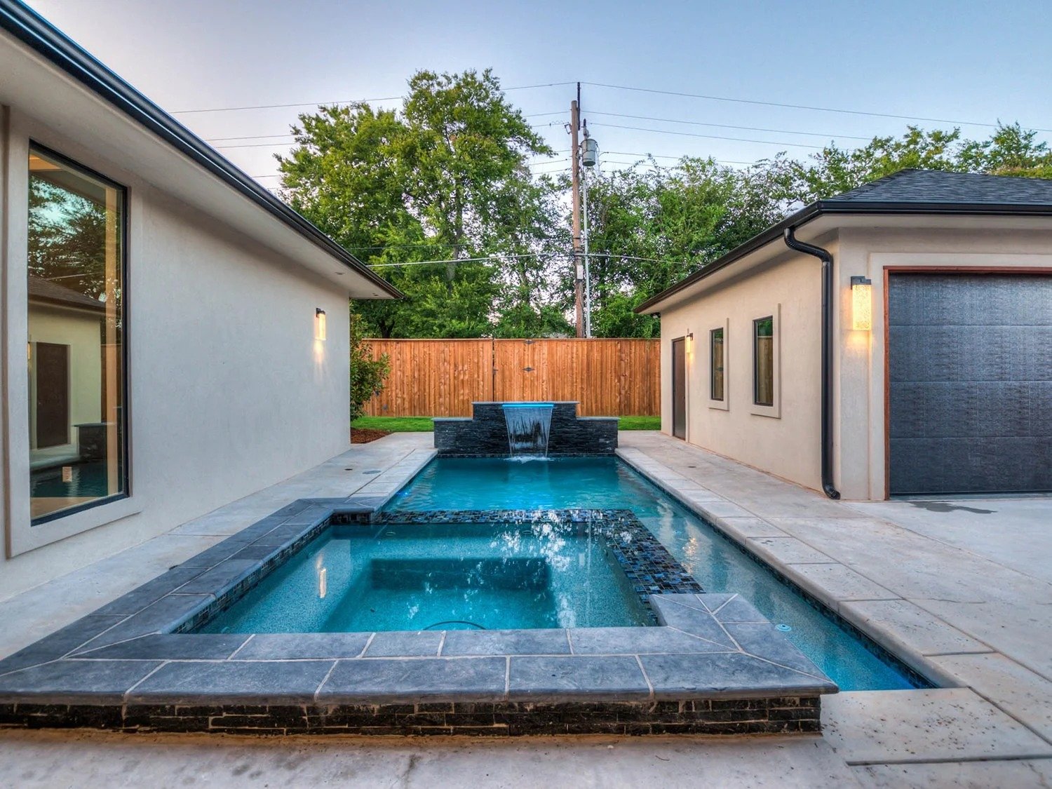 Backyard with swimming pool and waterfall feature, surrounded by concrete deck, between two modern houses, with a wooden fence and trees in the background.