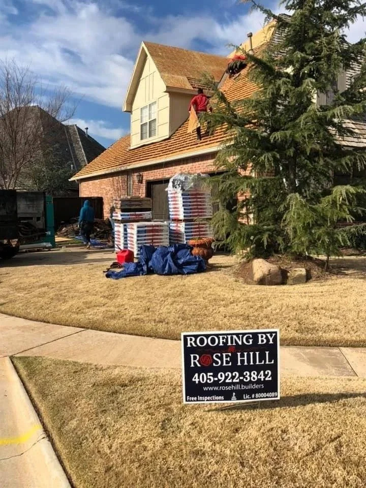 A house undergoing roofing work with workers on the roof and standing nearby, stacks of roofing materials in front, and a sign that says "Roofing by Rose Hill" in the front yard.