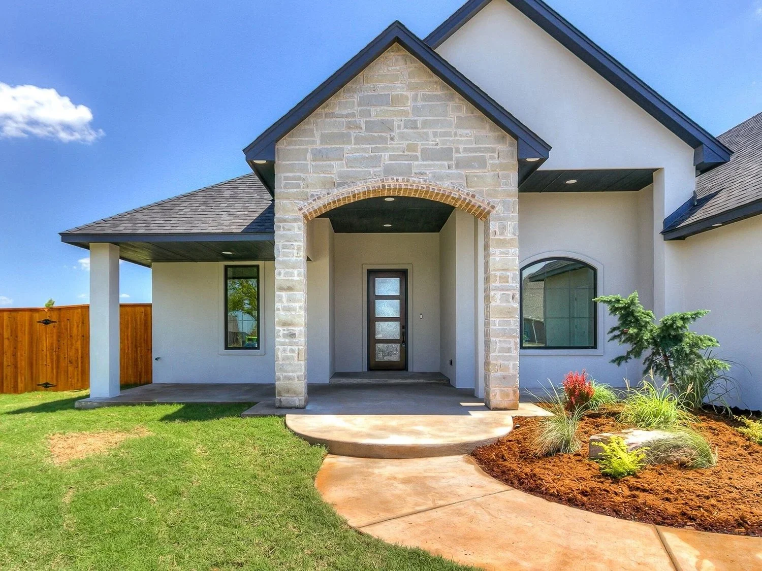 Front view of a modern house with a stone and stucco facade, a covered porch, a wooden fence, and a landscaped yard with grass, shrubs, and a small tree.