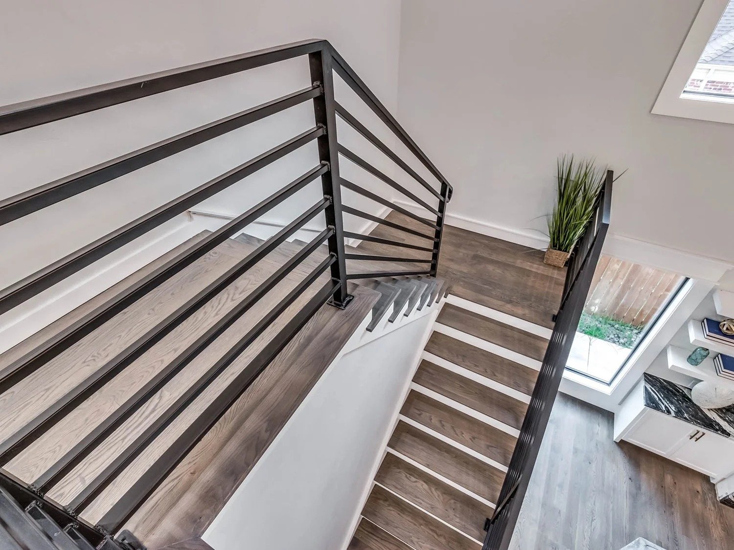 View from the top of a staircase with a black metal railing, showing wooden steps, a small landing with a potted plant, a large window, and part of a living space with shelves and a black marble table.