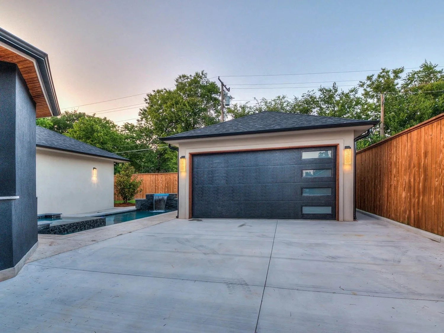 Modern backyard with a concrete patio, a pool area with a water feature, and a detached garage with a sleek black door and frosted glass windows, surrounded by a wooden privacy fence and green trees in the background.