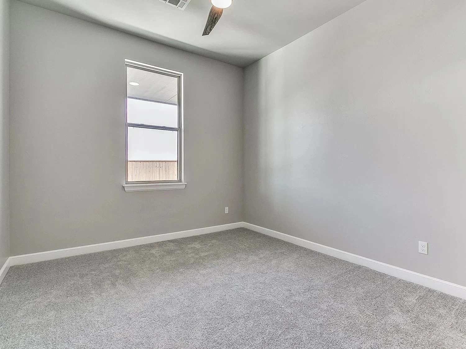 Empty room with gray walls, gray carpet flooring, a window with white trim, white baseboards, a ceiling fan, and an air vent.