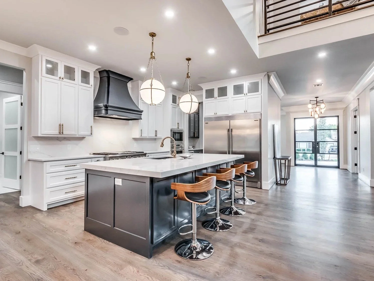 Modern kitchen with white cabinetry, black range hood, and a large island with dark gray base and white countertop. Four wooden barstools are lined up at the island. Stainless steel appliances, including a double-door refrigerator and built-in oven, 