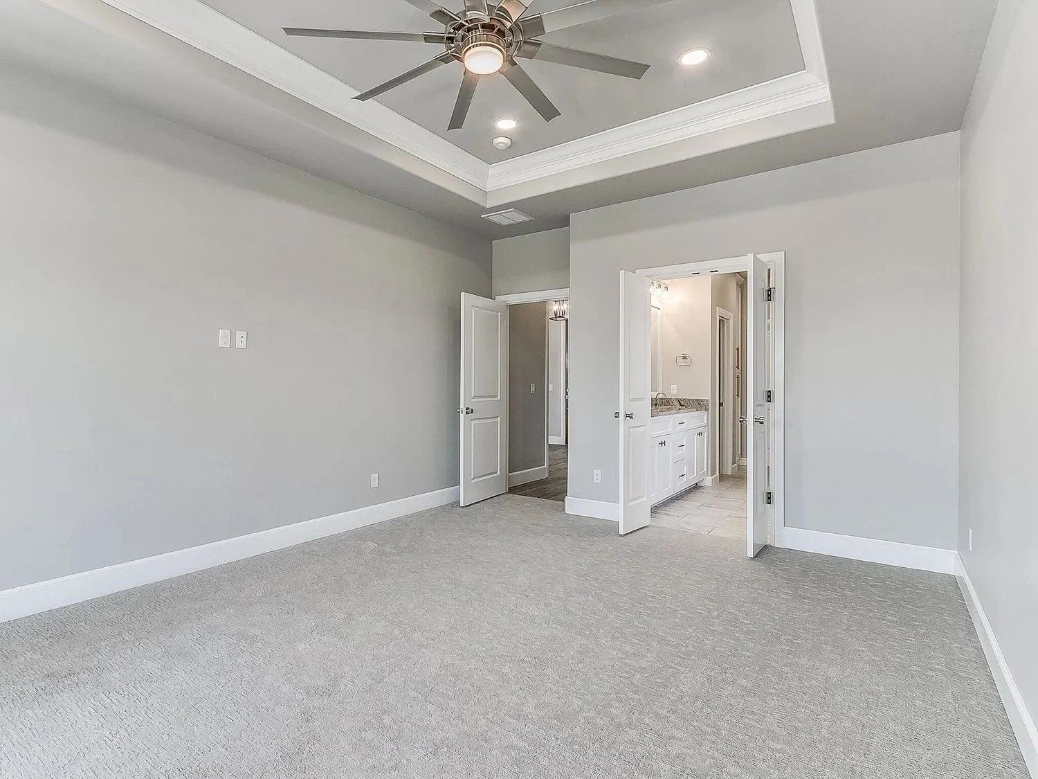Empty room with light gray walls, white baseboards, beige carpet, and a tray ceiling with a modern ceiling fan and recessed lighting. Open doors reveal a bathroom vanity and a hallway.