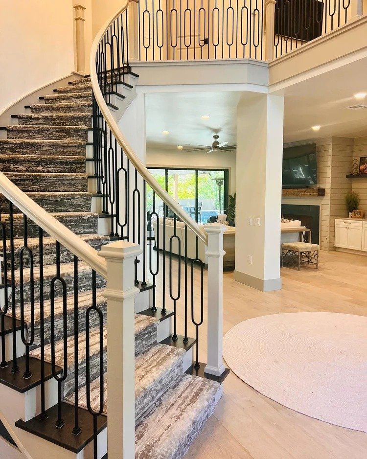 Interior view of a home with a curved staircase featuring black metal railings and a carpeted runner, leading to an upper level, with a living room area in the background including a TV, ceiling fan, and sliding glass doors opening to an outdoor area
