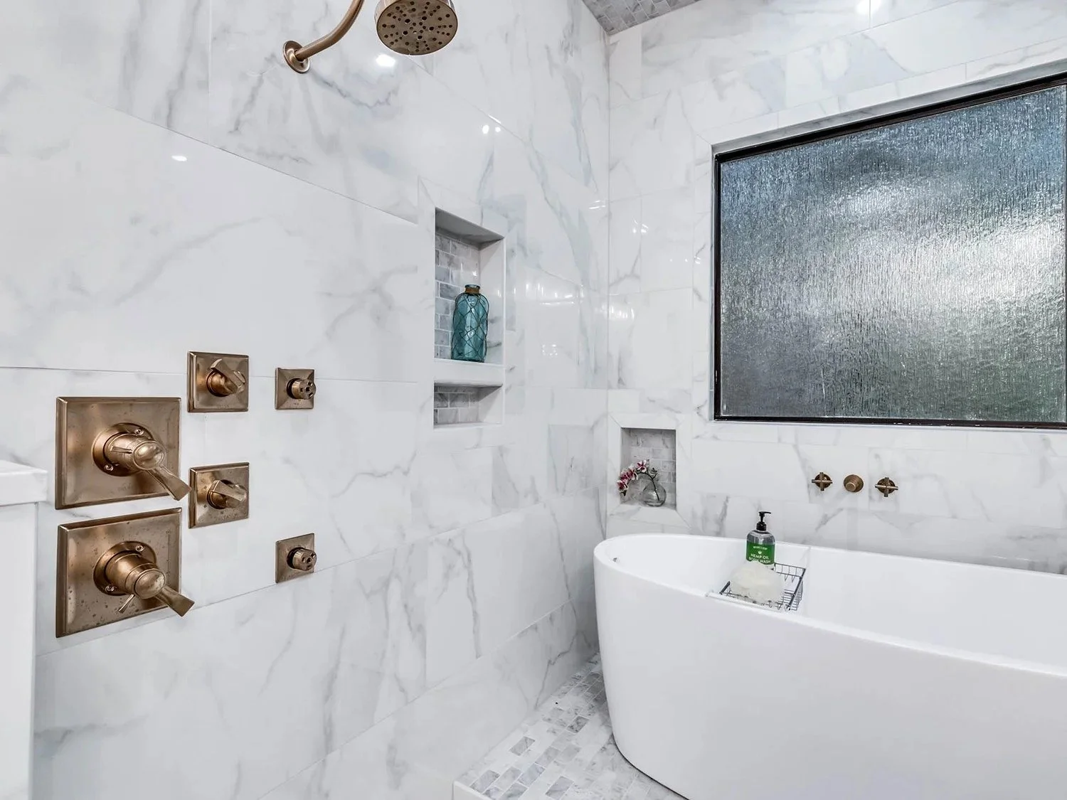 Modern bathroom with marble walls, a large frosted window, a white bathtub, a showerhead, and brass fixtures.