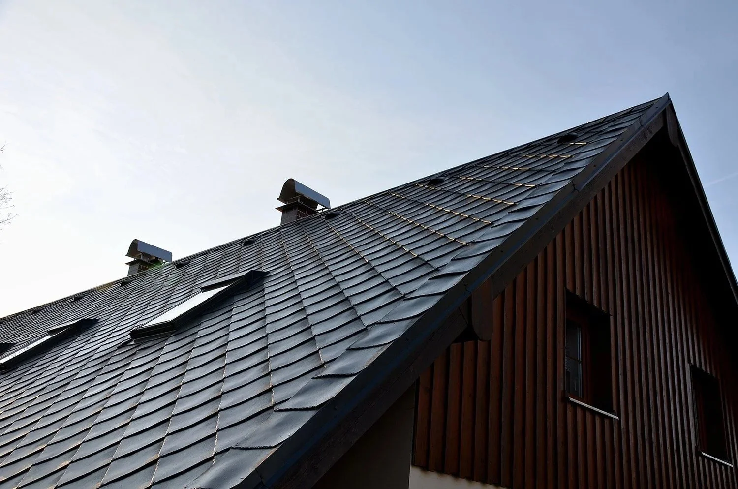 Close-up of a house roof with dark gray shingles, side of the building with wooden vertical siding, and windows, against a clear sky.