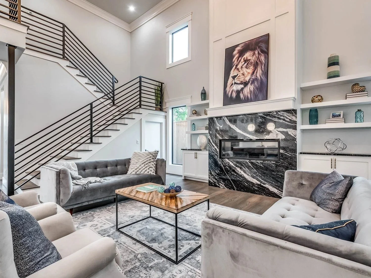 Living room with modern design, featuring a black and white marble fireplace, a large lion portrait above, built-in white shelves with decorative vases and books, a staircase with black metal railing, and light-colored sofas with pillows. There's a w