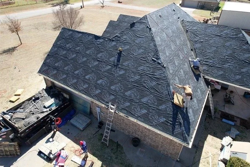 Aerial view of a house with a black tarp covering the roof during roofing work, with workers installing or repairing the roofing.