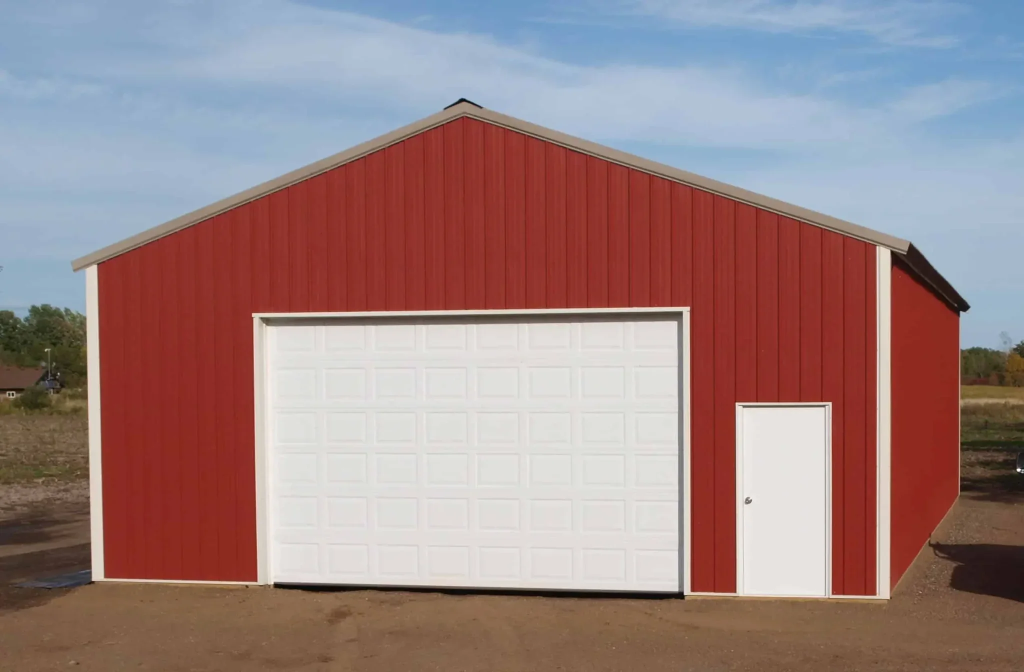 A red barn-style building with a white garage door and a small side door, situated outdoors on a dirt surface under a blue sky with some clouds.