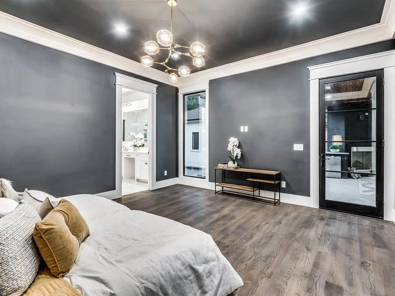 A modern living room with dark gray walls, white window and door trim, hardwood floors, and a black door with glass panels. There is a white sofa with beige pillows, a black and wood console table with white orchids, and a contemporary light fixture 