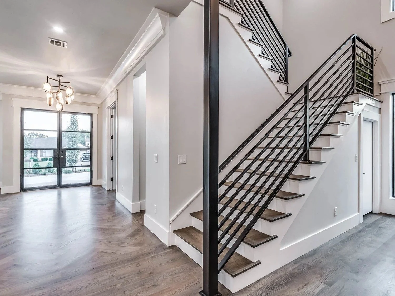 Modern interior of a house with a staircase, hardwood floors, and large windows with a view of a yard and neighboring houses.