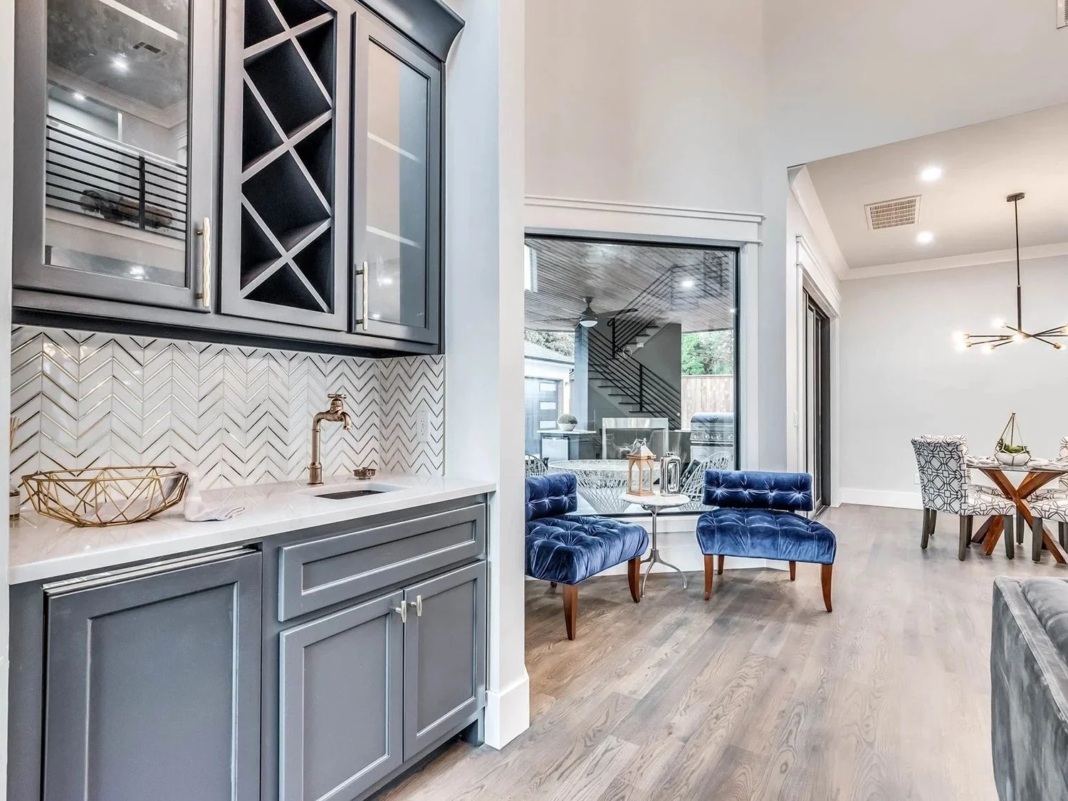 Living room with blue velvet chairs, a glass sliding door, a wooden dining table with patterned chairs, and kitchen cabinets with a backsplash.