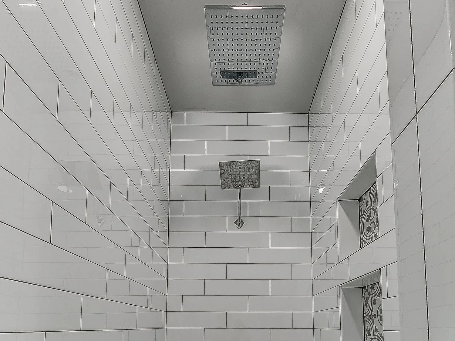 A shower area with white tiled walls, a ceiling-mounted showerhead, a square rain showerhead, and decorative tiles in a niche.