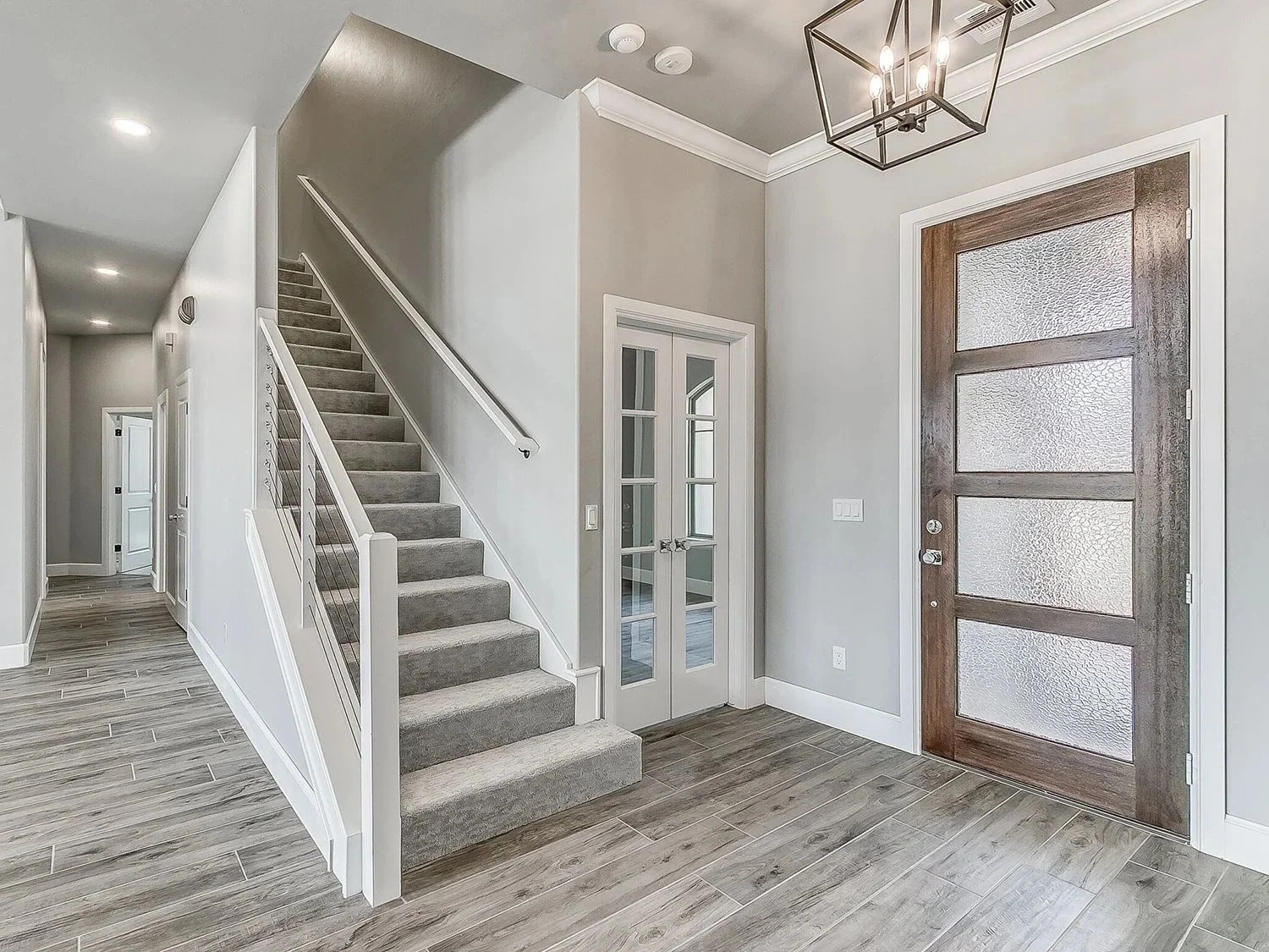 Interior view of a modern house entryway with a front door featuring textured glass panels, a small glass-paneled door, a staircase with gray carpet, and wood-look tile flooring.