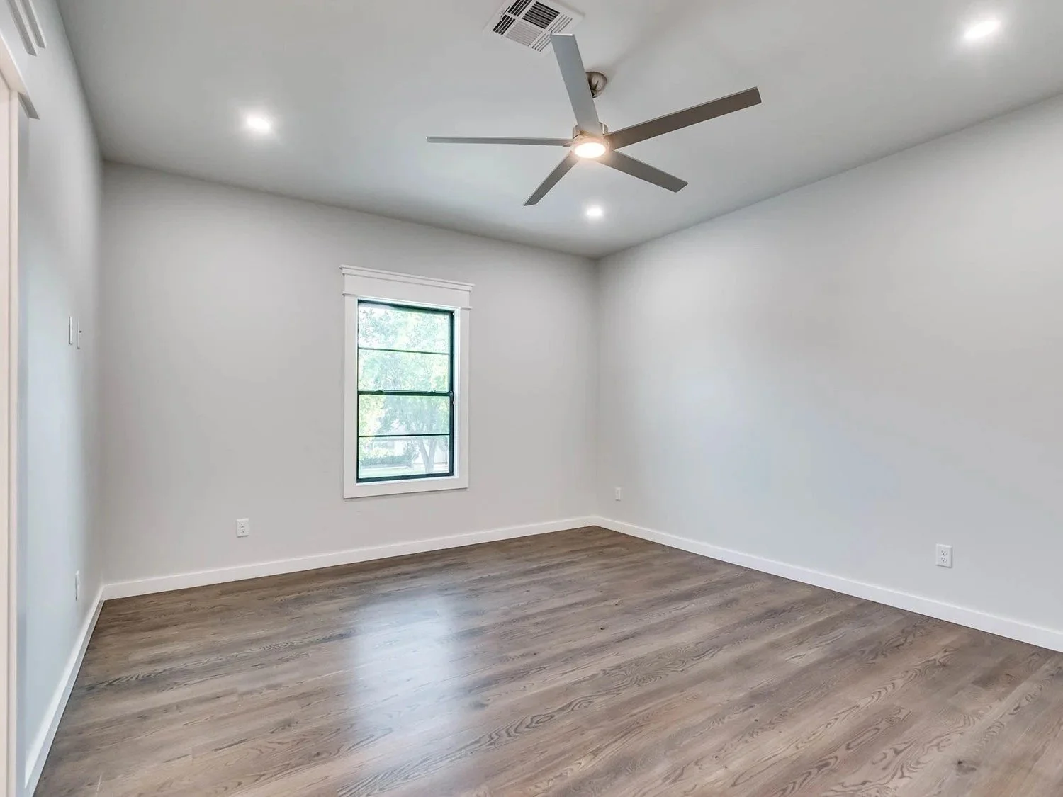 Empty room with white walls, wooden floor, one window, ceiling fan, and ceiling lights.