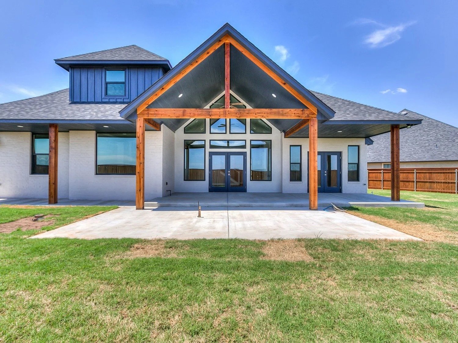 Newly constructed house with a large front porch and patio, featuring wooden beams and a gabled roof, with grass and a wooden fence in the yard, under a clear blue sky.