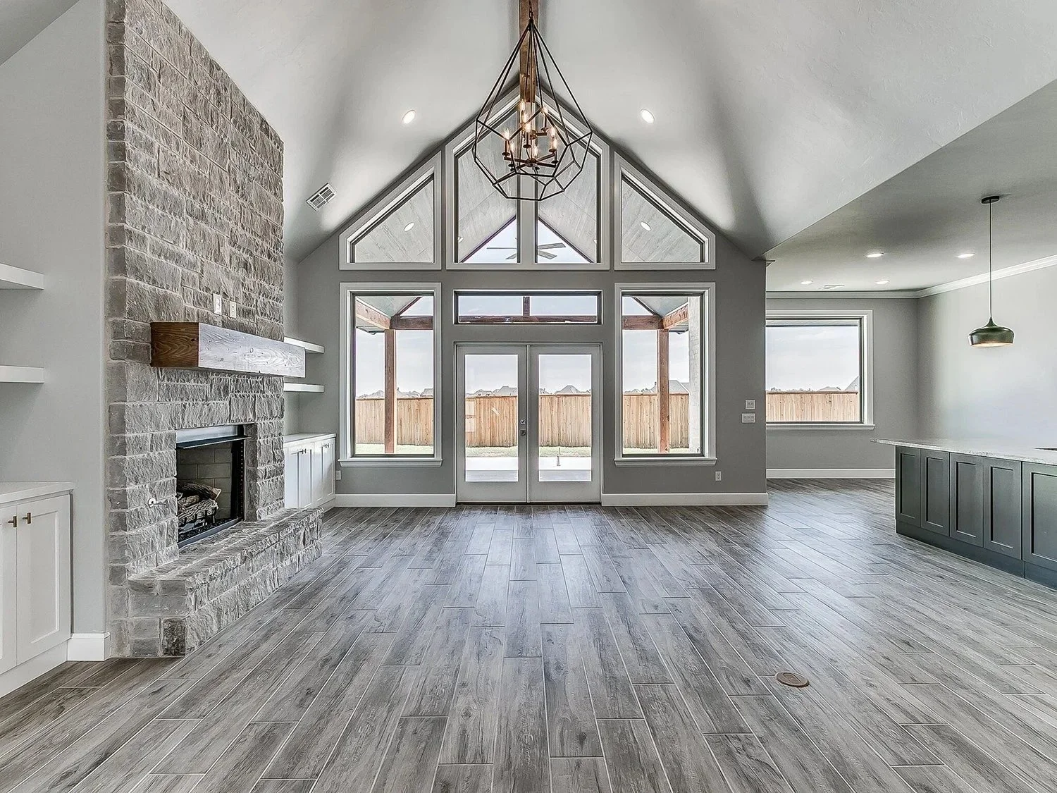 Empty living room with a stone fireplace, large window and door at the back, high vaulted ceiling with chandelier, and wood-look flooring.