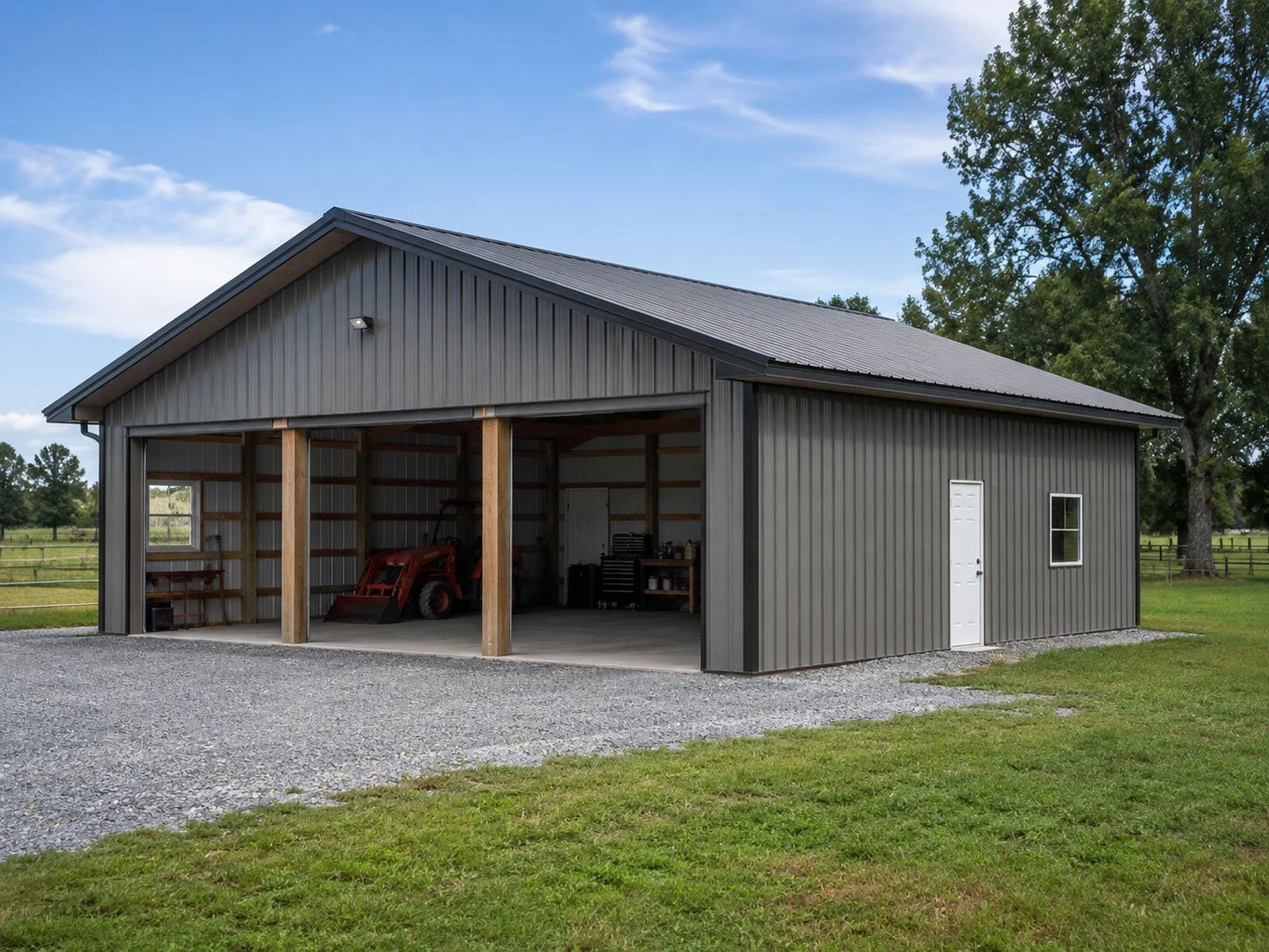 A metal barn with an open garage section showing a small tractor inside, and a white door and window on the side, situated on a grassy area with trees and a fence in the background.