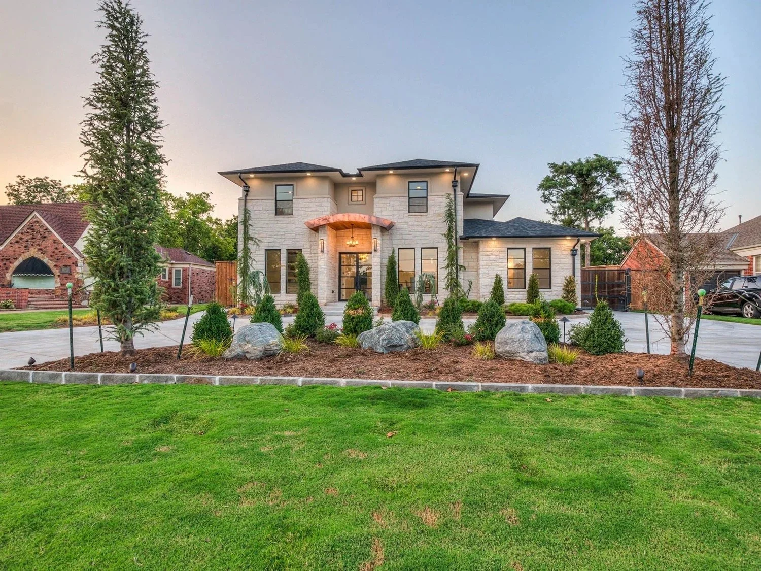 A modern two-story house with a landscaped front yard featuring green grass, small shrubs, and large rocks. The house has light-colored brick exterior, black-framed windows, and a decorative front porch with a chandelier, illuminated by exterior ligh