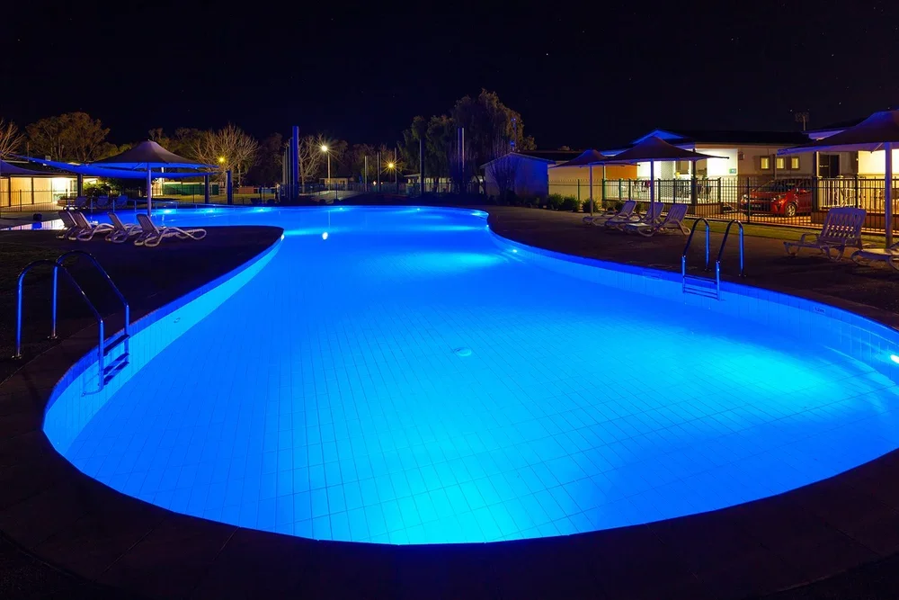 Nighttime view of an illuminated outdoor swimming pool with lounge chairs, umbrellas, and surrounding residential buildings.