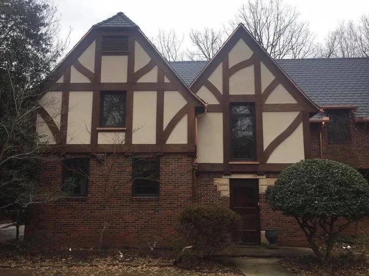 Front view of a house with Tudor-style architecture, brick lower level, and white upper sections with brown timber detailing. Trees and shrubs are in the yard.