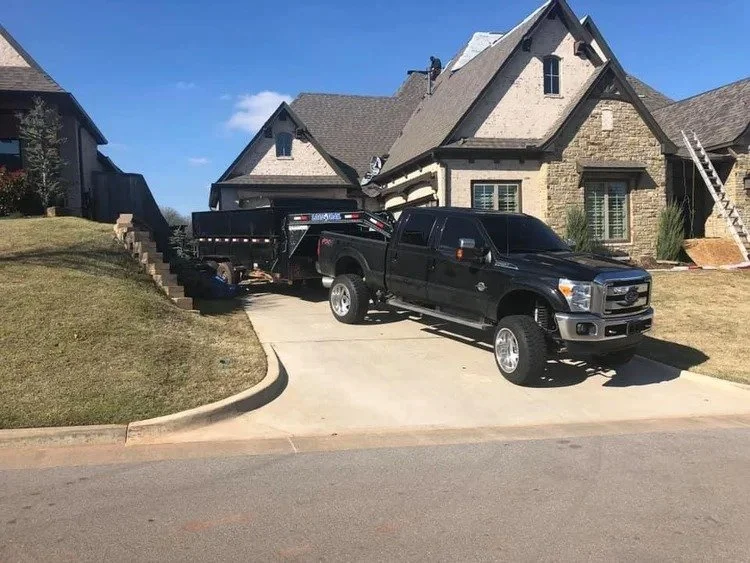 A black pickup truck parked on a driveway next to a house with a steep roof. The truck is towing a flatbed trailer.