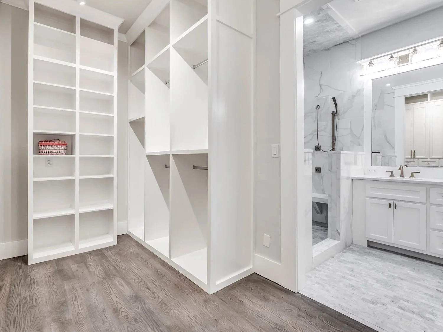 Empty walk-in closet with white shelving and a view into a bathroom with a marble shower and vanity