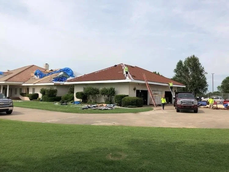 Workers repairing the roof of a house, with several vehicles parked nearby and building materials on the lawn.